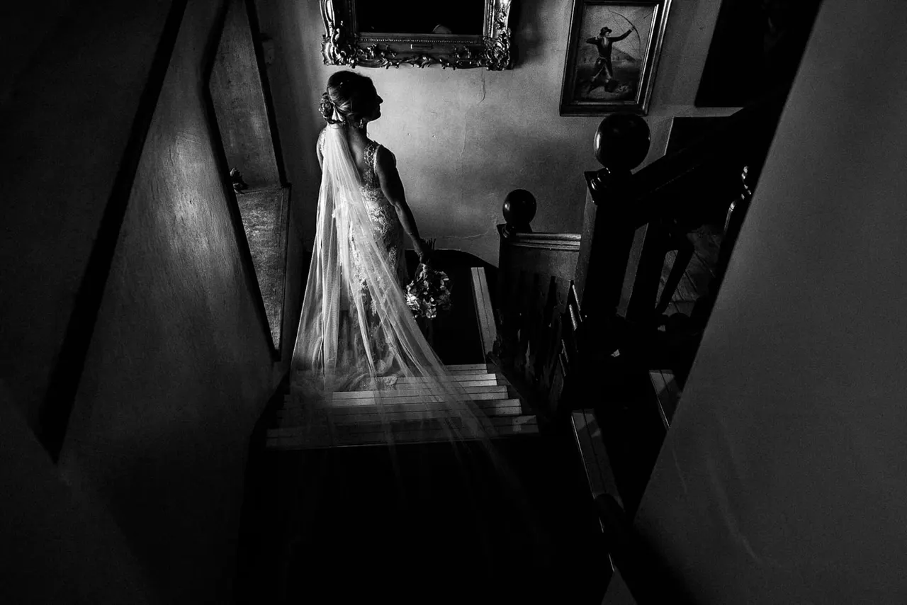 Bride in white gown and veil standing in a narrow period interior with attendants, photographed from above in black and white.