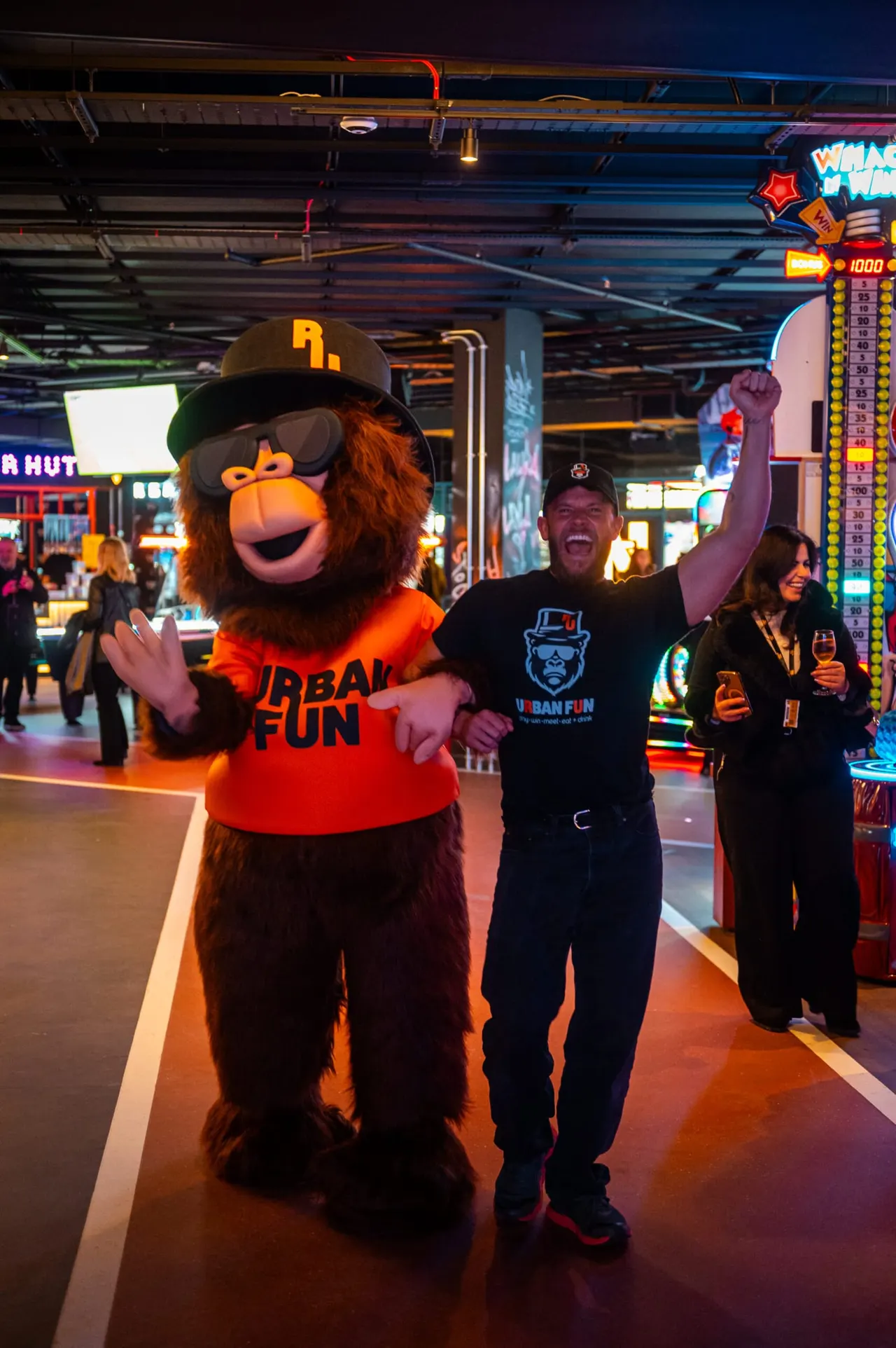 A mascot character in red and brown costume poses with a smiling man and woman inside a brightly lit indoor entertainment arcade.