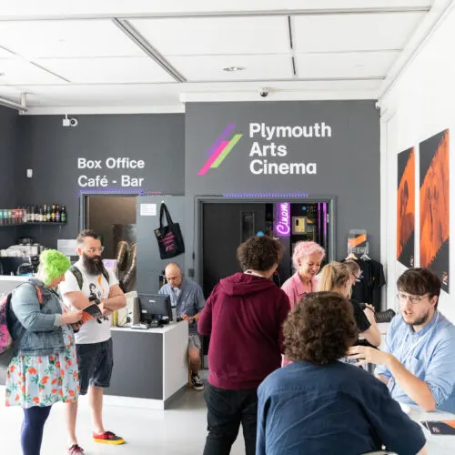 People queue and socialise at a café bar counter inside a cinema foyer.