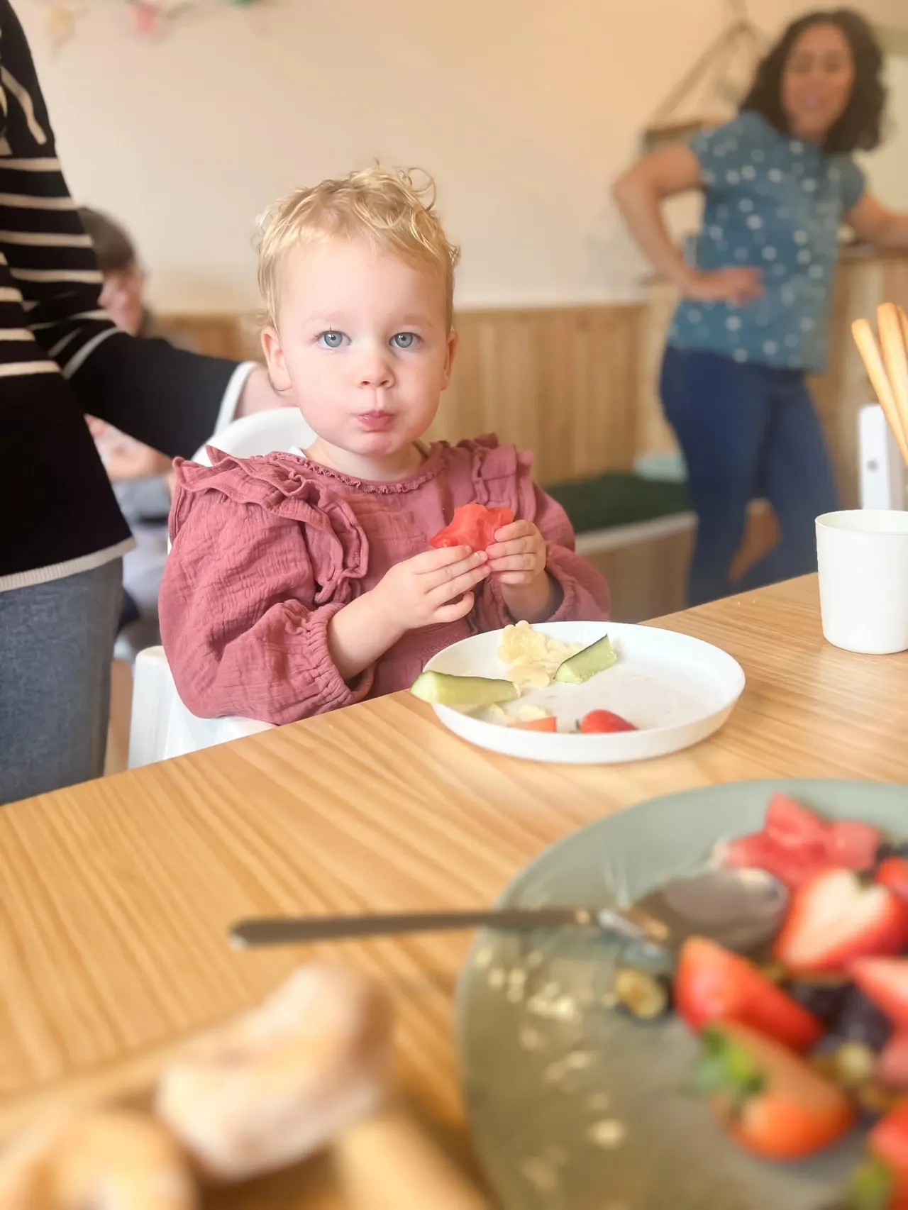 Young toddler in pink top eating food at a table during a soft play session with other children in the background.