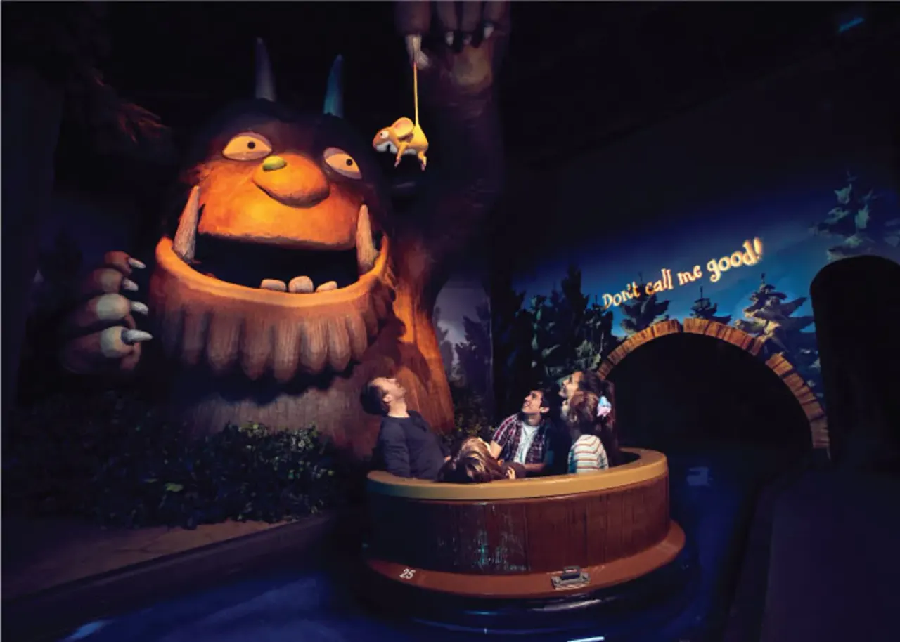 Visitors in a boat ride encounter a large animatronic Gruffalo character with glowing orange features in a darkened themed attraction.