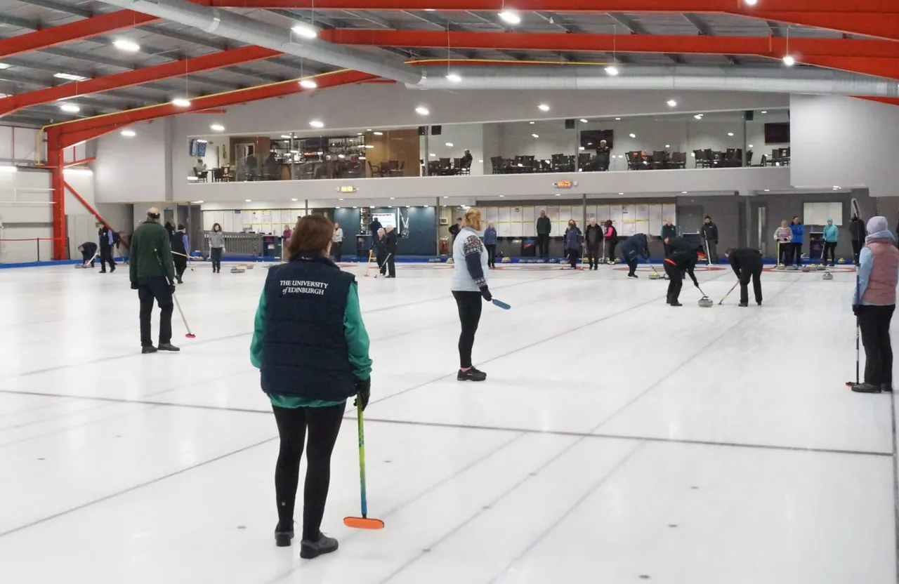 Multiple people playing curling on indoor ice rinks within a modern sports facility with red roof beams.