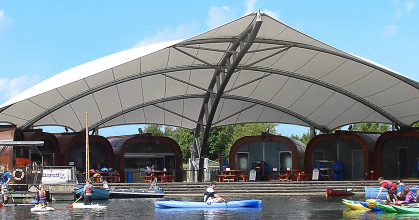 Families paddling kayaks and stand-up paddleboards on a broad, shallow river under a large fabric canopy shelter.