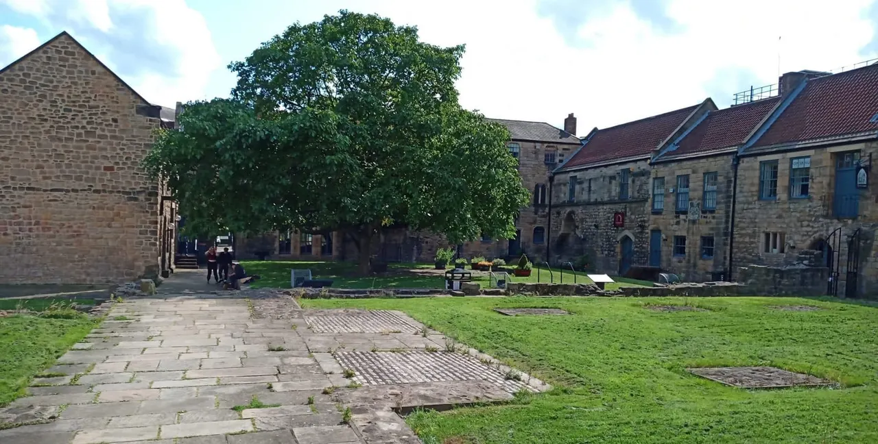 Stone-built courtyard with green lawn, paved pathways, a large tree, and historic stone buildings surrounding the space.