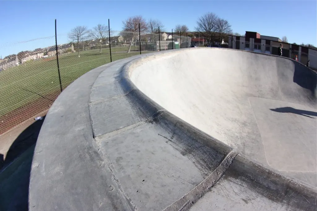 Empty concrete skate bowl with curved ramps, fencing and spectator area visible under clear sky.