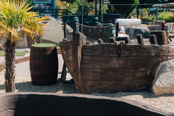 Rustic wooden barrel and ship-themed props at a mini golf course with palm trees and gravel landscaping.