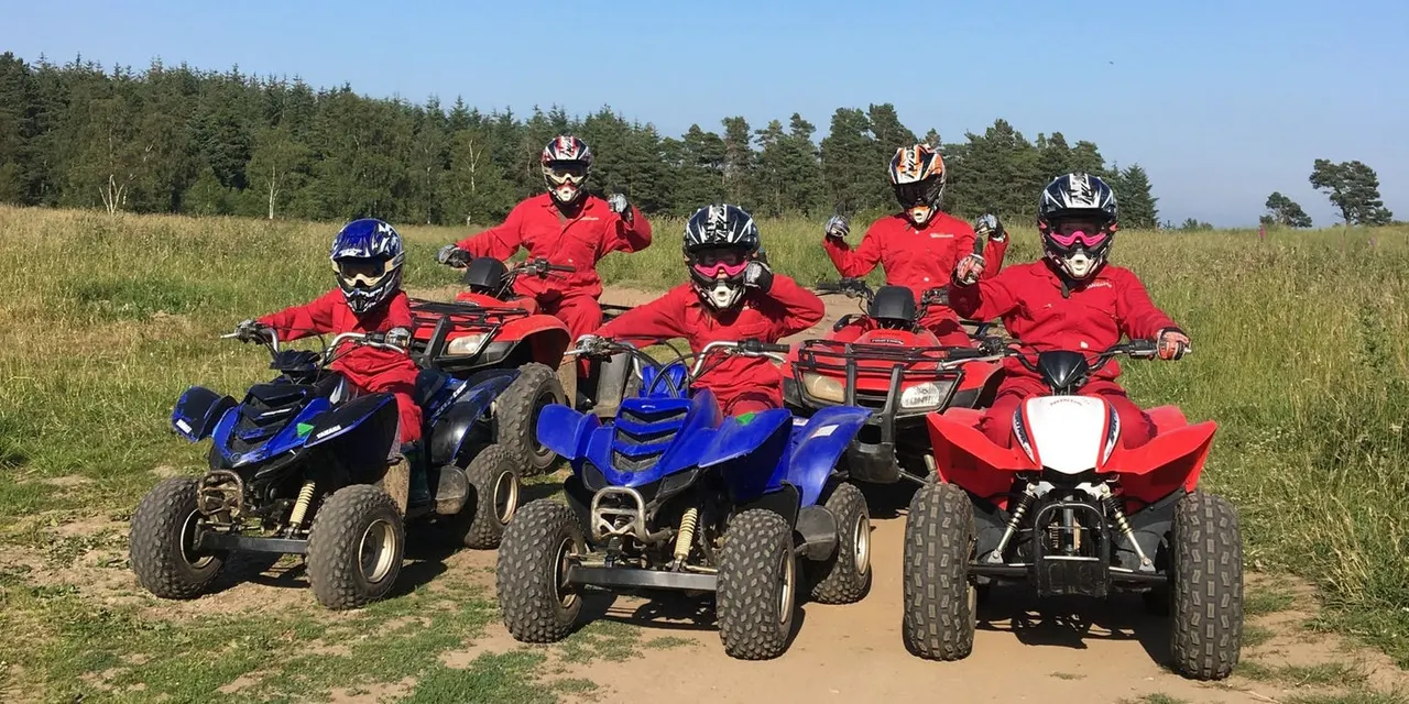 Group of five riders in red outfits on blue and red quad bikes lined up on a dirt track with woodland and fields behind.