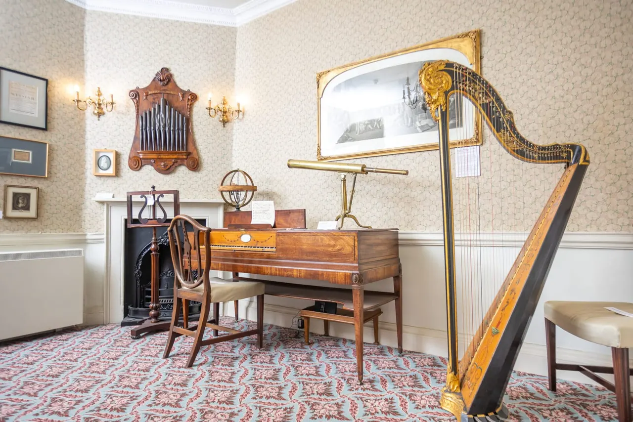 A music room displaying antique wooden instruments including a harp, organ pipes and a telescope, with period furniture and decorative wall sconces.