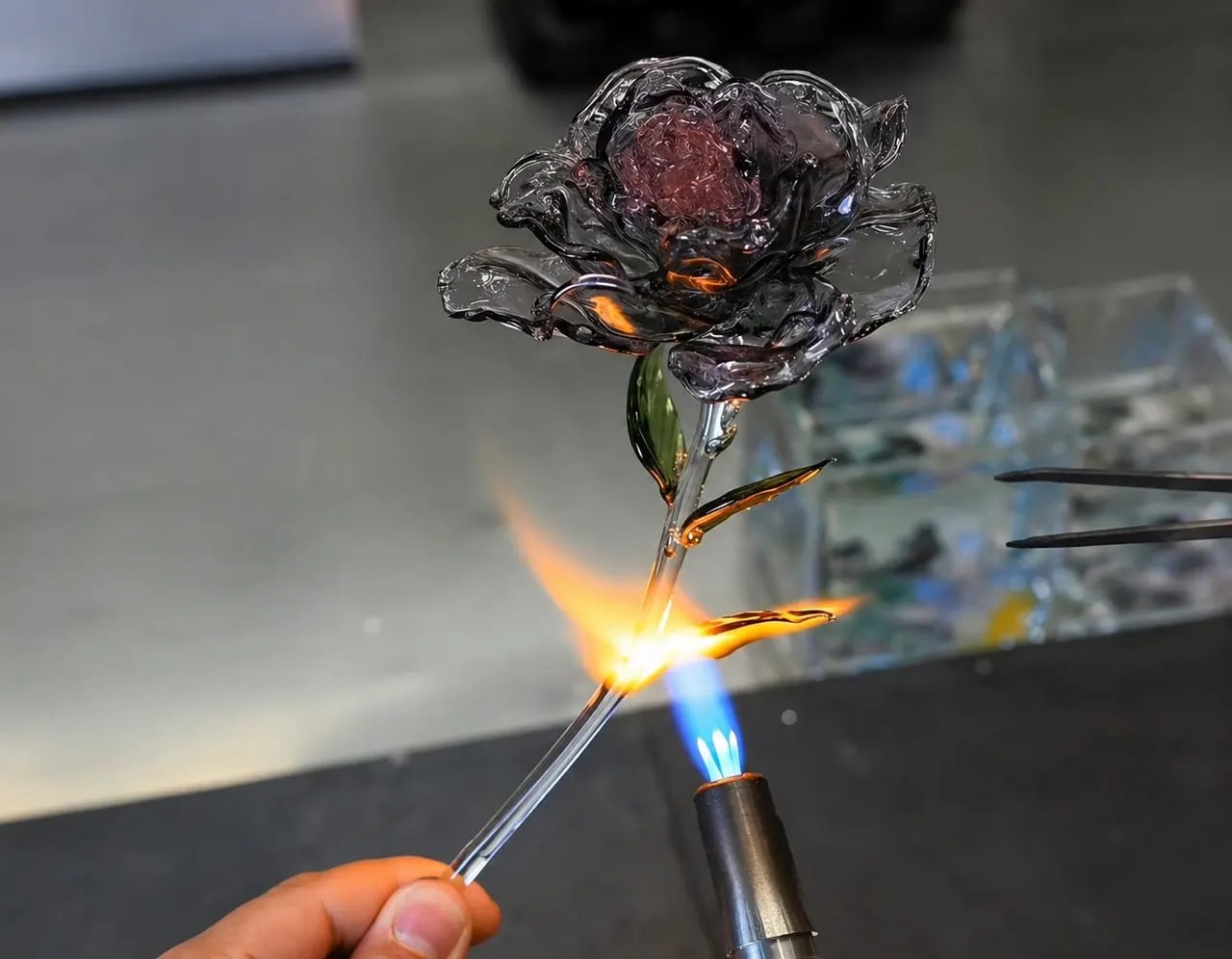 A glassblower's hand holds a torch to a molten glass flower with pink petals and green stem during creation.