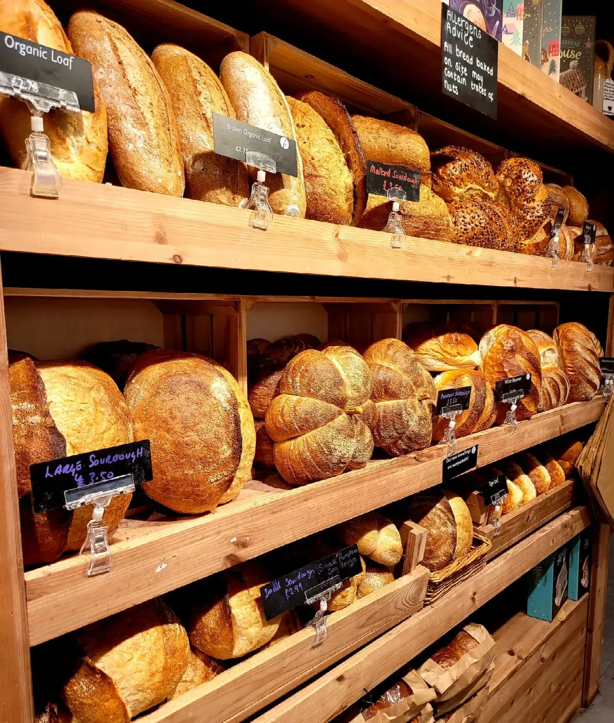 Wooden shelves displaying various freshly baked loaves and rolls with handwritten price labels.