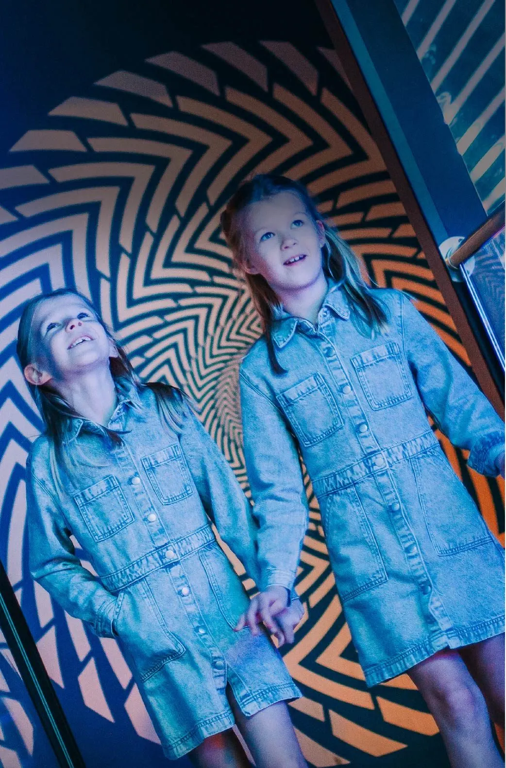 Two young children wearing denim shirts gaze upward in front of an illuminated geometric optical illusion display.