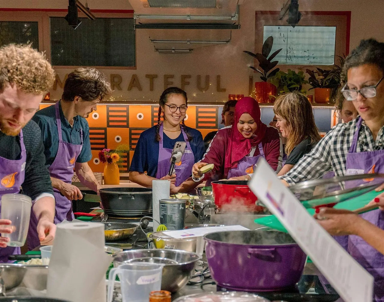 A group of people wearing purple aprons gather around a cooking station, preparing food together in a bright kitchen classroom.