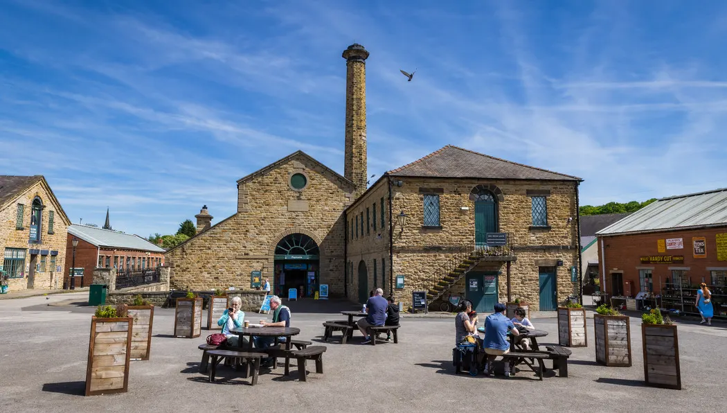 A historic stone mill complex with tall chimney, visitors seated at picnic tables in a sunny courtyard.