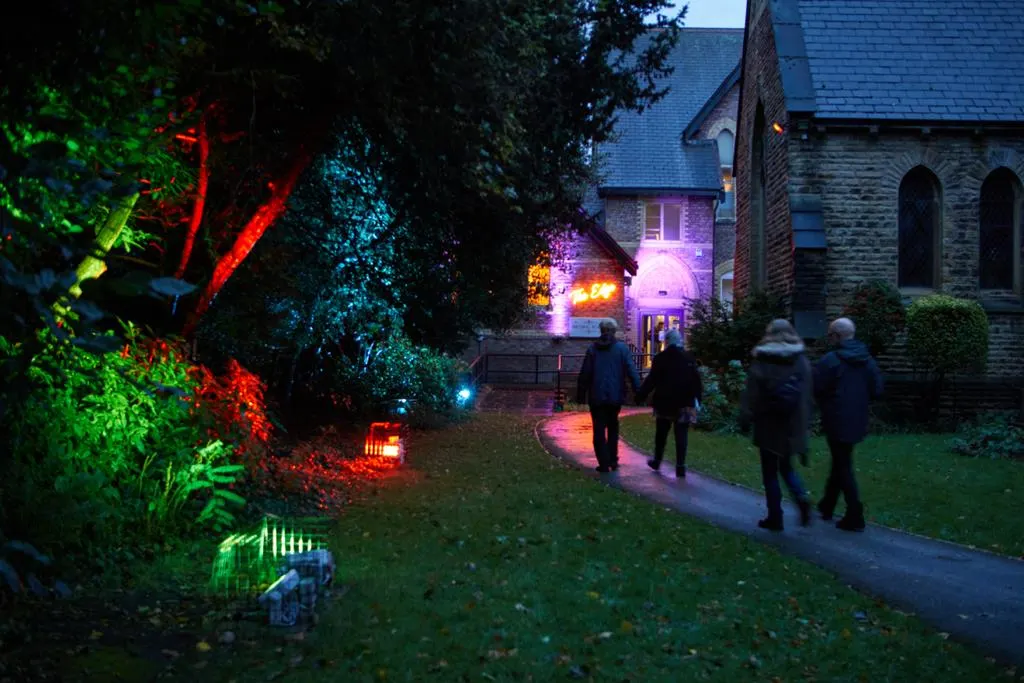 Visitors walk through an illuminated garden towards stone buildings at dusk, with coloured uplighting on trees and pathways.