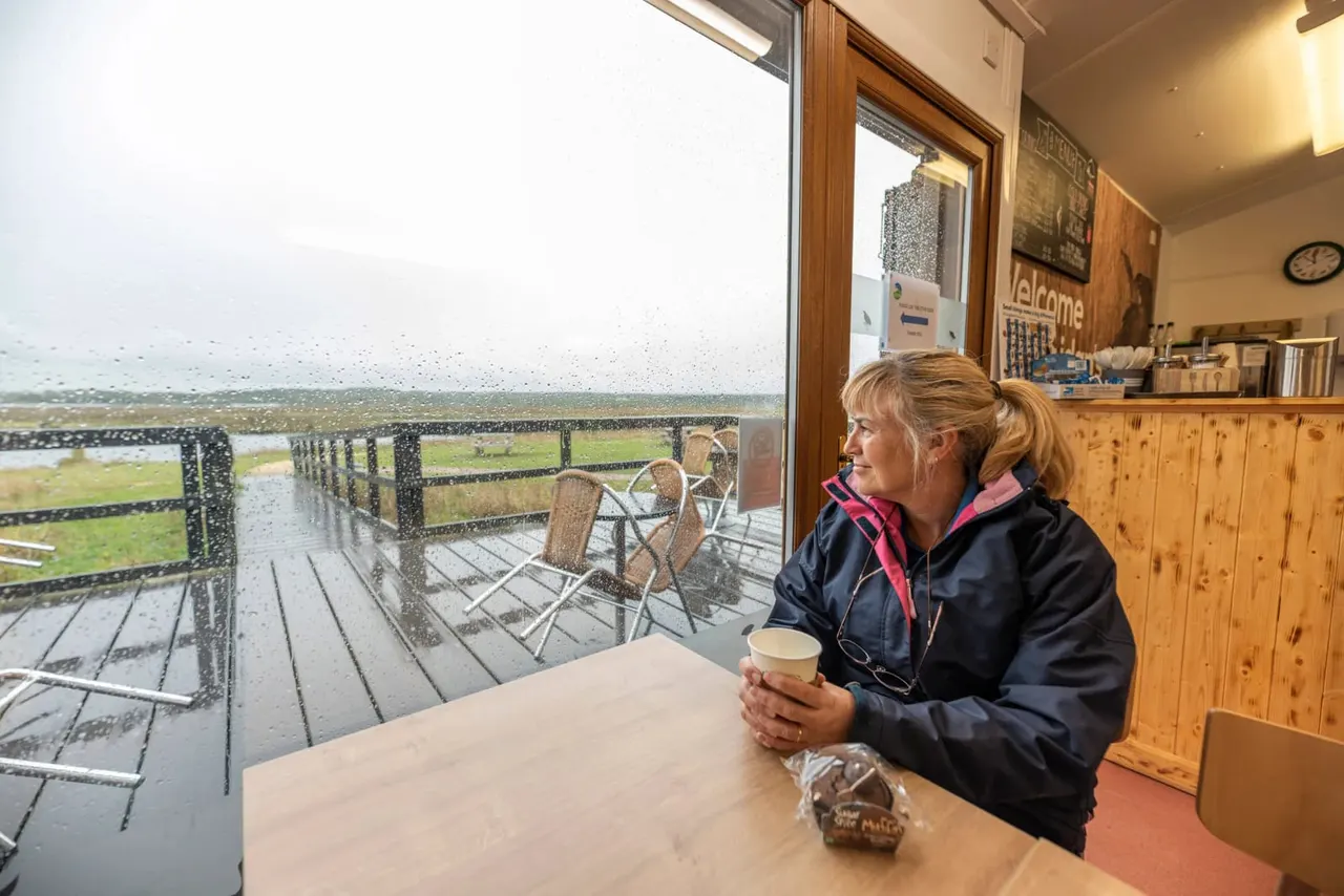 Woman with hot drink looking out at wetland habitat with water, reeds, and distant birds from indoor viewing room.