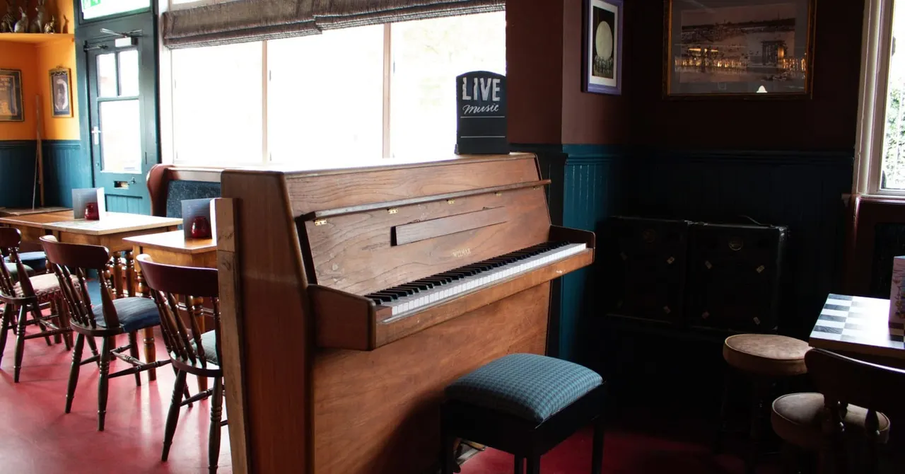 Interior of a pub with a wooden upright piano in the foreground and seating areas with wooden chairs and tables visible in the background.