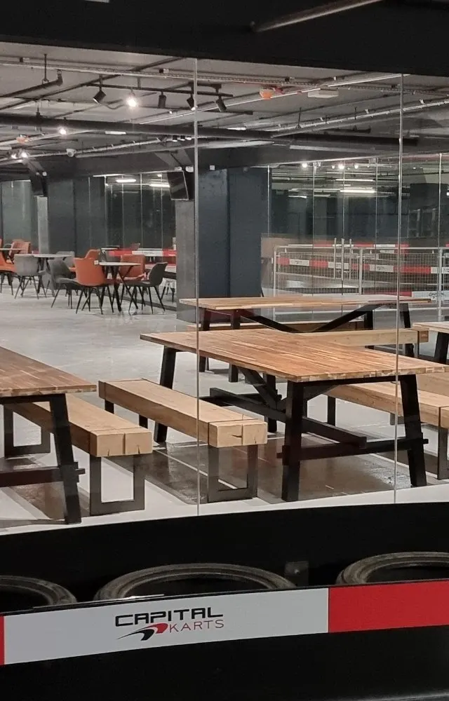 Modern indoor leisure facility with wooden picnic tables and benches in foreground, glass partitions, and orange seating areas visible in background.