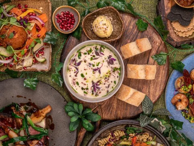 Overhead view of an assorted spread of cooked dishes, breads and fresh herbs arranged on wooden boards and plates.