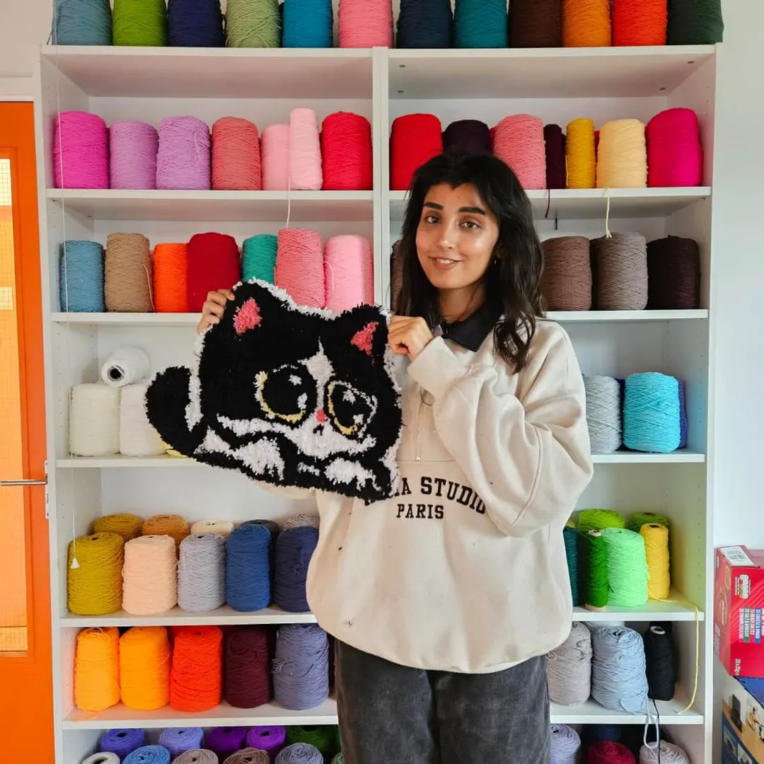 Woman holding a tufted black cat cushion in front of shelves stocked with colourful yarn rolls.