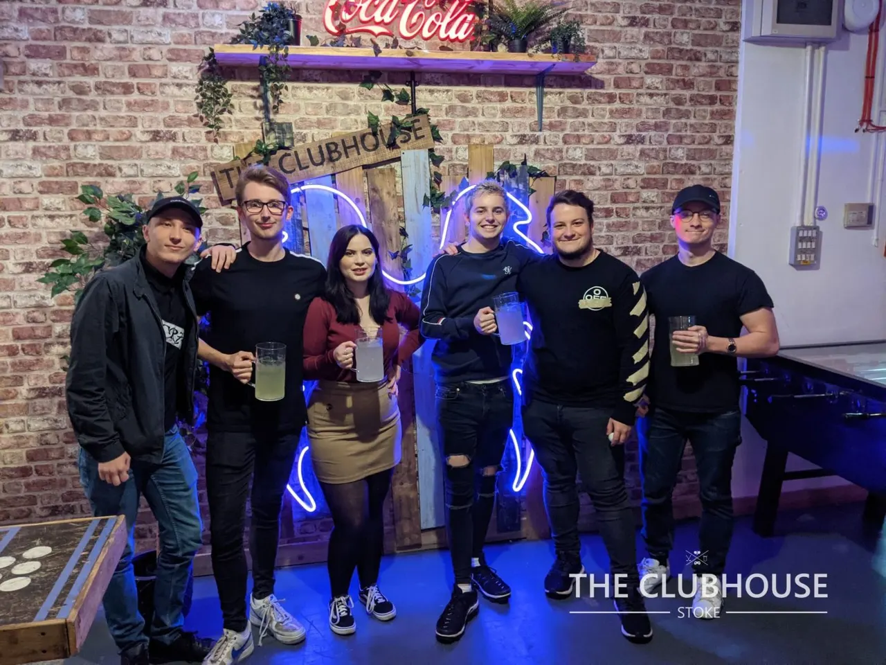 Six young people stand together holding drinks in a venue with exposed brick walls, blue neon lighting, and a Coca-Cola sign.