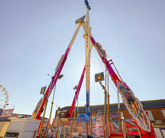 A tall fairground ride with red arms and a central wooden pole against a clear sky, with brick buildings and other attractions visible around it.