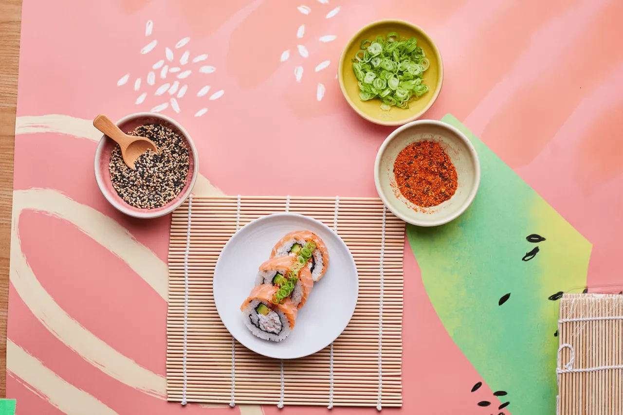 Sushi roll with salmon and green onions surrounded by sesame seeds, spiced powder, and sliced spring onions on a pink background.