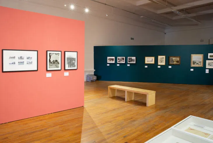 Gallery space with framed artworks displayed on coral and teal walls, wooden bench seating, and wooden flooring.