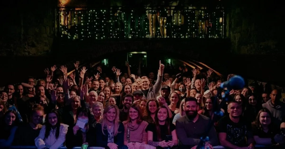 Large crowd of people with raised hands enjoying a live event in an illuminated underground venue.