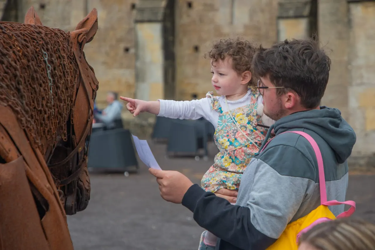 An adult holding a small child points at a brown horse in a castle courtyard setting.