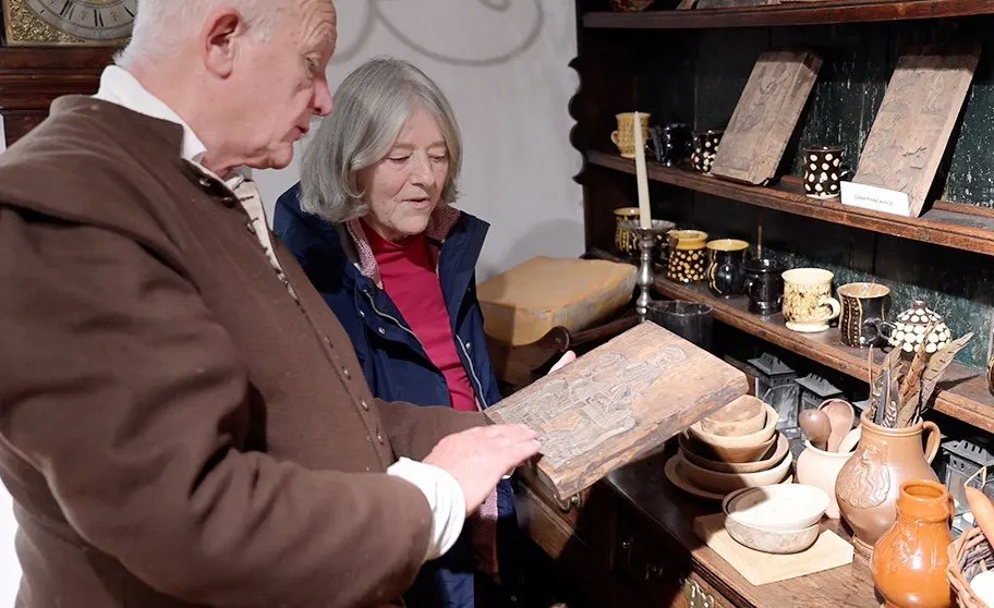 Two older visitors examining a wooden artefact in a period room display with pottery and household items.