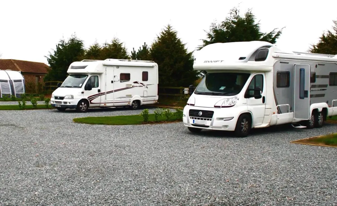 Three white motorhomes parked on gravel at a campsite with evergreen trees and a cottage in the background.
