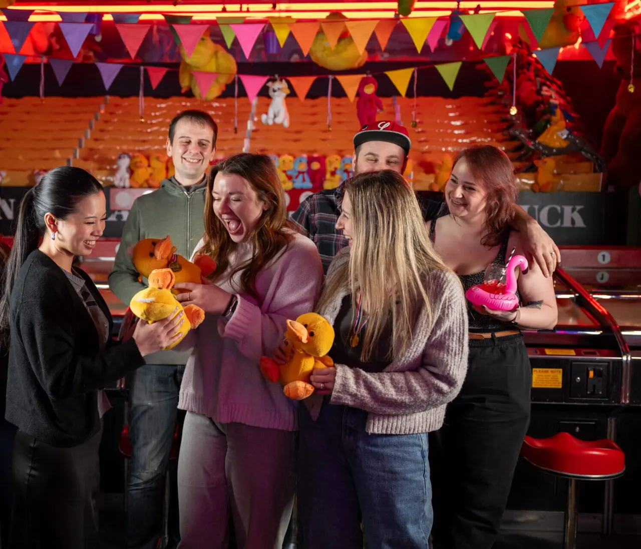 Group of young adults laughing whilst holding won stuffed toys in a colourful arcade bar with neon lighting.