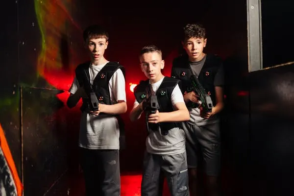 Three young people holding laser guns in a dimly lit arena with red lighting effects.