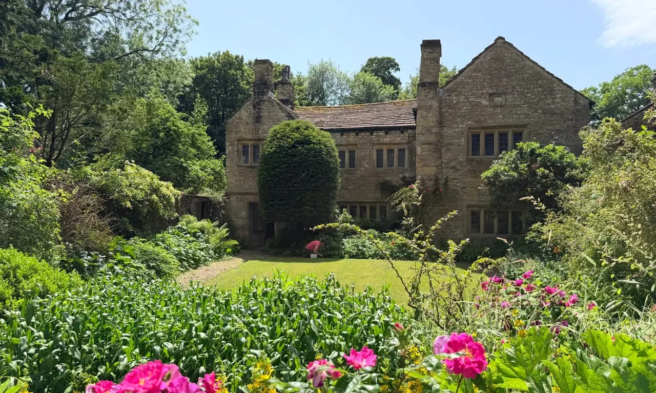 Stone historic building surrounded by mature trees, with manicured gardens featuring pink flowering plants and green lawns in the foreground.