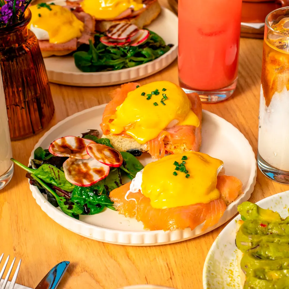 Eggs Benedict with smoked salmon and hollandaise sauce served on white plates with green salad and colourful drinks.