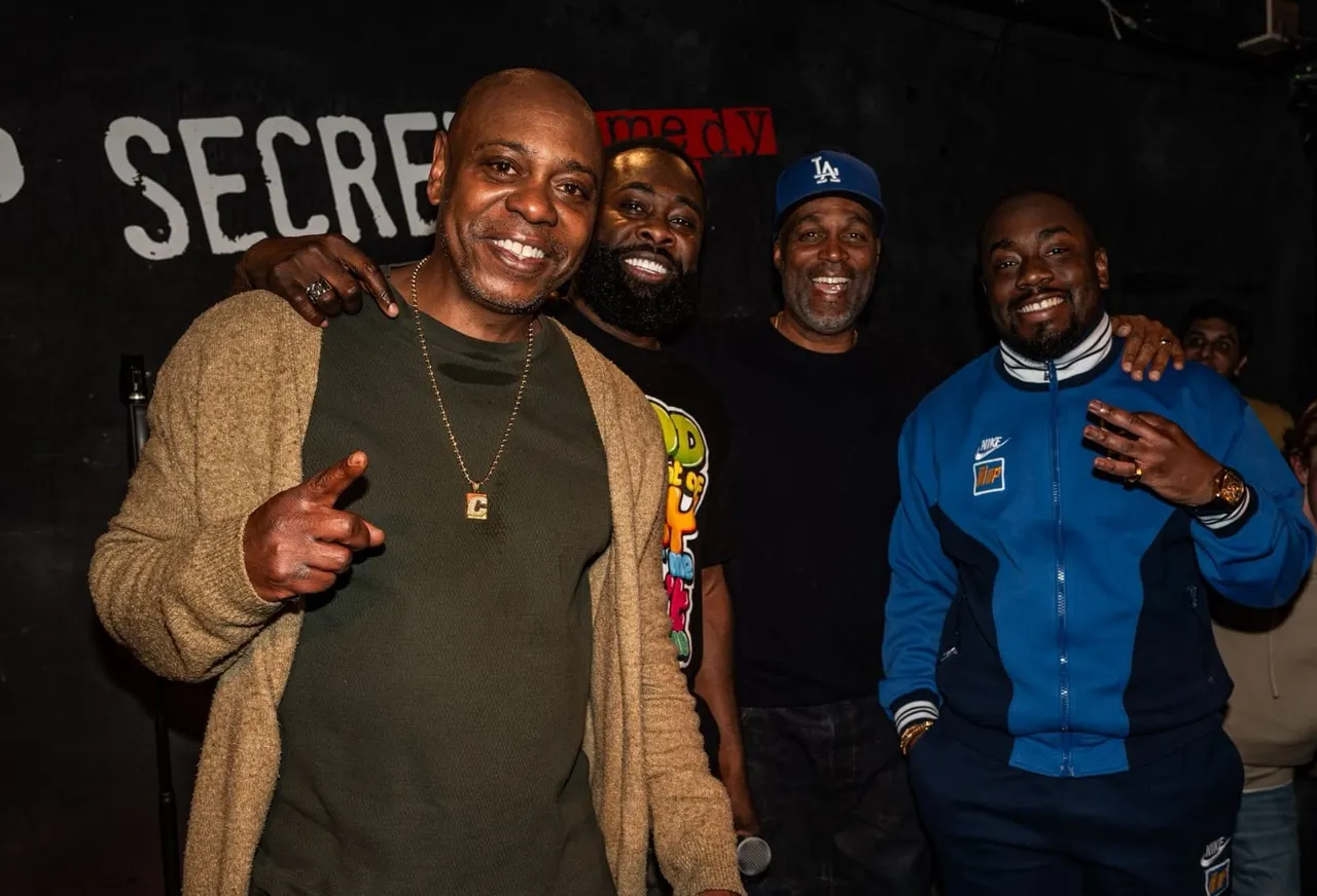 Four men smiling at a comedy club event, with partial signage visible in the background.