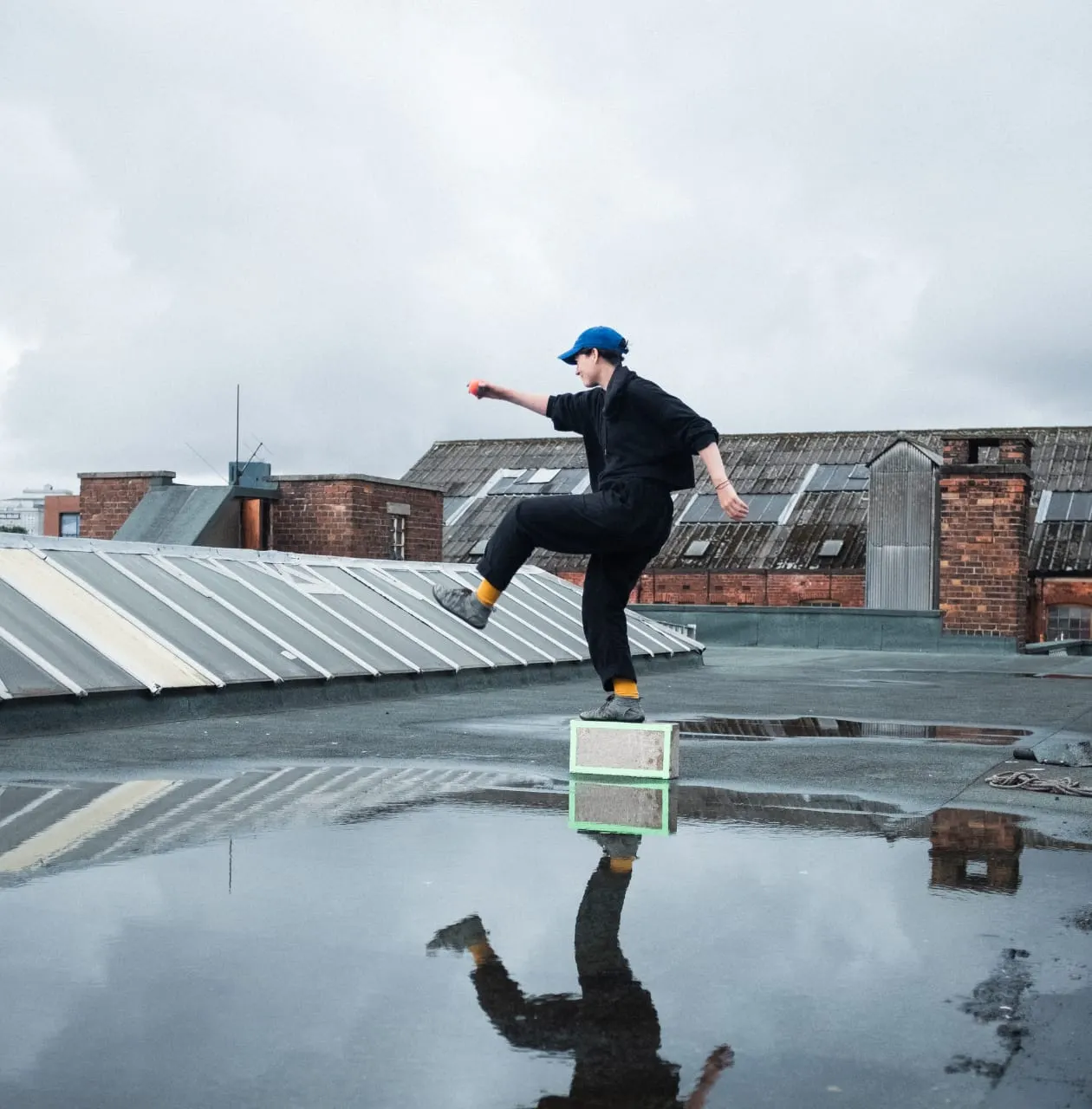 Person in blue cap and black clothing performing parkour jump over green box on wet rooftop with brick buildings behind.
