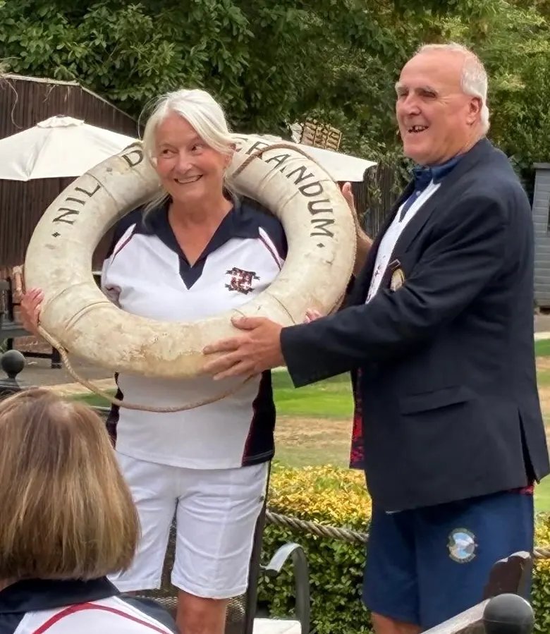 Two adults holding a large decorative life ring at an outdoor leisure venue, with spectators and garden setting visible.