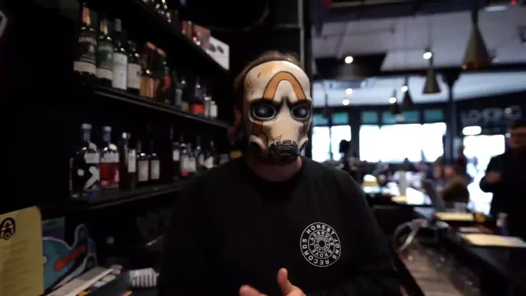 Person wearing a skeletal mask and dark clothing inside a bar with shelves of bottles behind them.