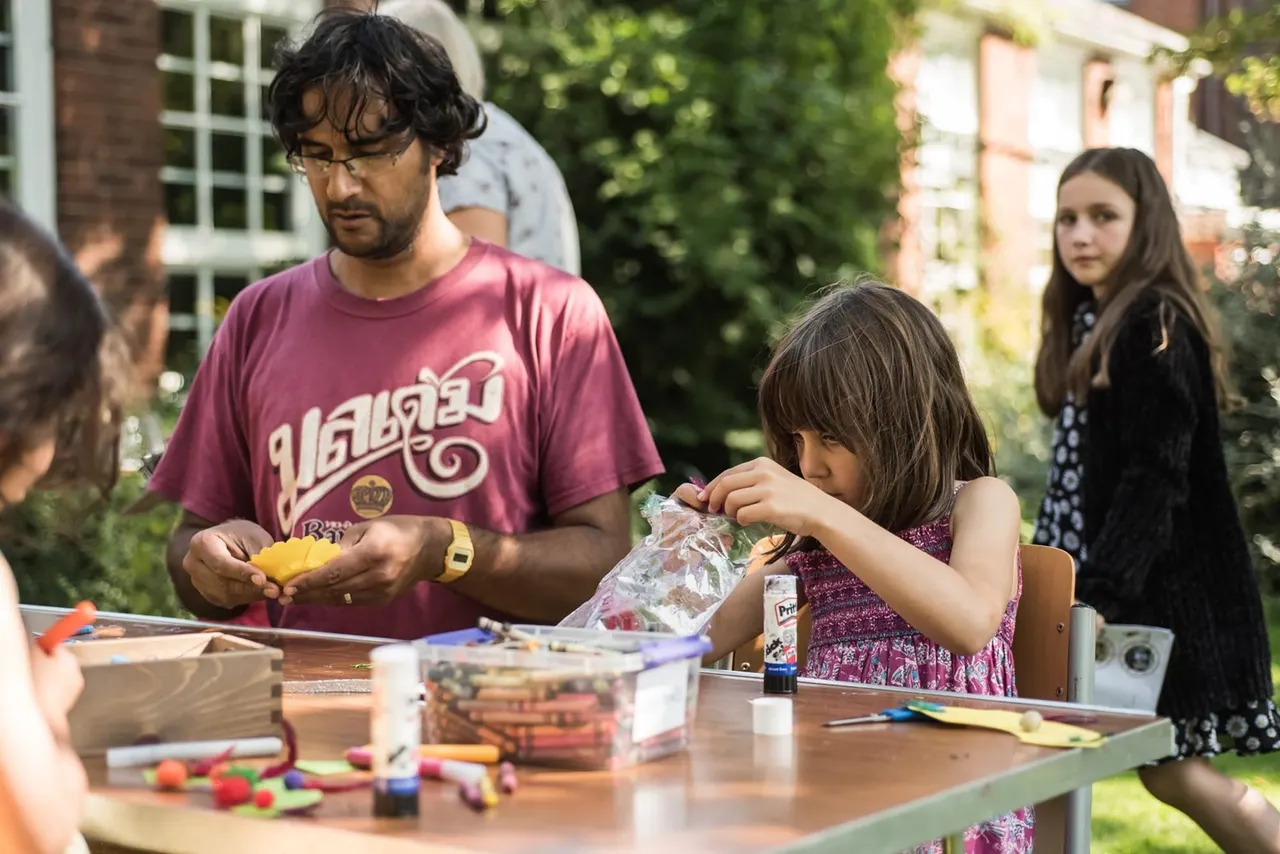 An adult and children participate in a craft activity at an outdoor table in a sunny garden setting beside historic brick buildings.