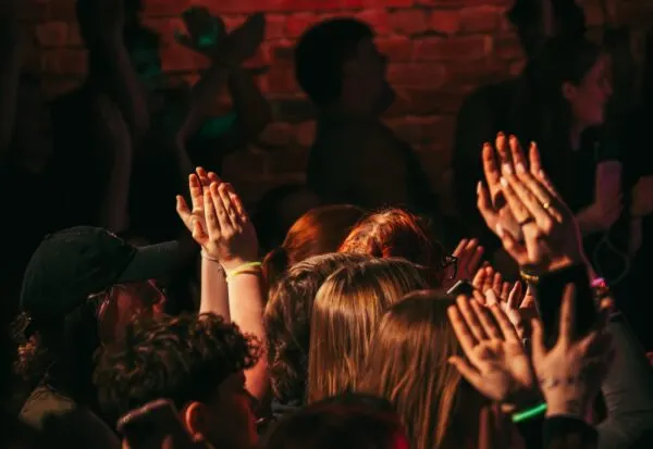 Crowd of people dancing and raising their hands at a live music event with red-lit brick wall backdrop.