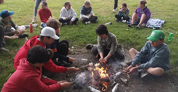 Group of children and adults gathered around a campfire on grass, roasting food and socialising outdoors.