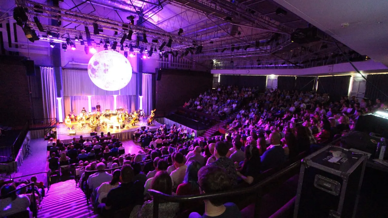 Large auditorium with hundreds of seated audience members watching an orchestra perform on an illuminated stage beneath a glowing moon projection.
