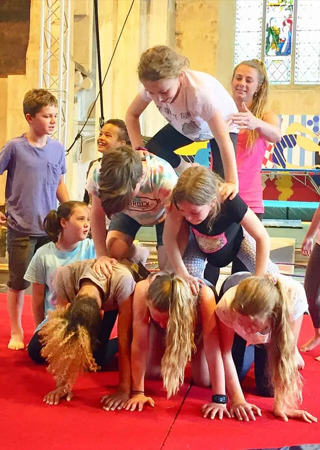 Children and an instructor engaging in a circus training activity on a red mat indoors.