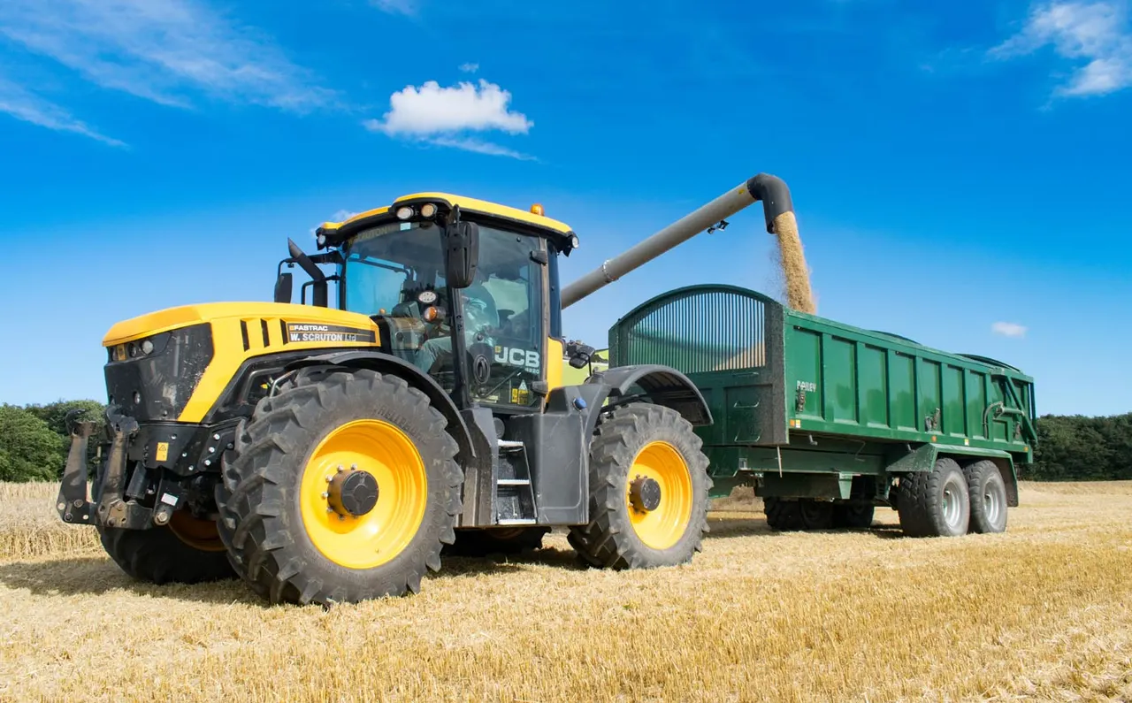 Yellow and black JCB tractor harvesting grain into a green trailer in a stubble field under blue sky.