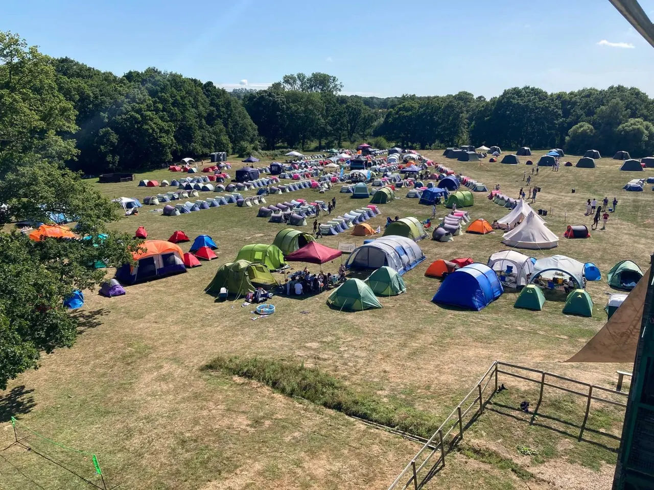 Large camping field with hundreds of colorful tents and bell tents pitched across grassland surrounded by woodland.
