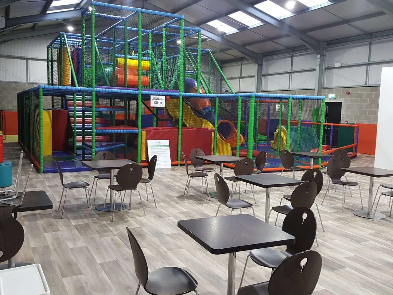 Indoor play facility with large coloured climbing structure, slides and netting, with dining tables and chairs in foreground.