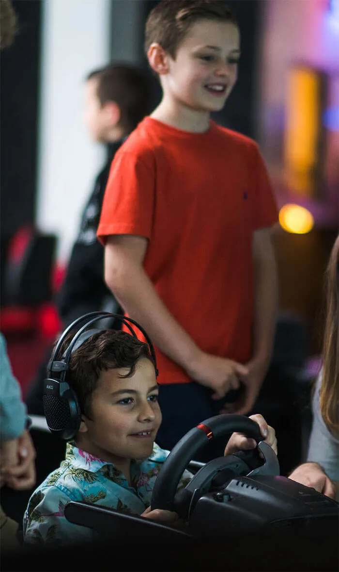 Child wearing headphones holding a gaming controller whilst another child in red stands nearby at an indoor entertainment venue.