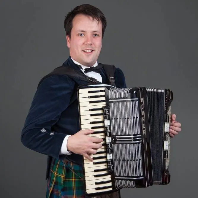 Man in formal dark jacket and tartan kilt holding an accordion against a grey background.