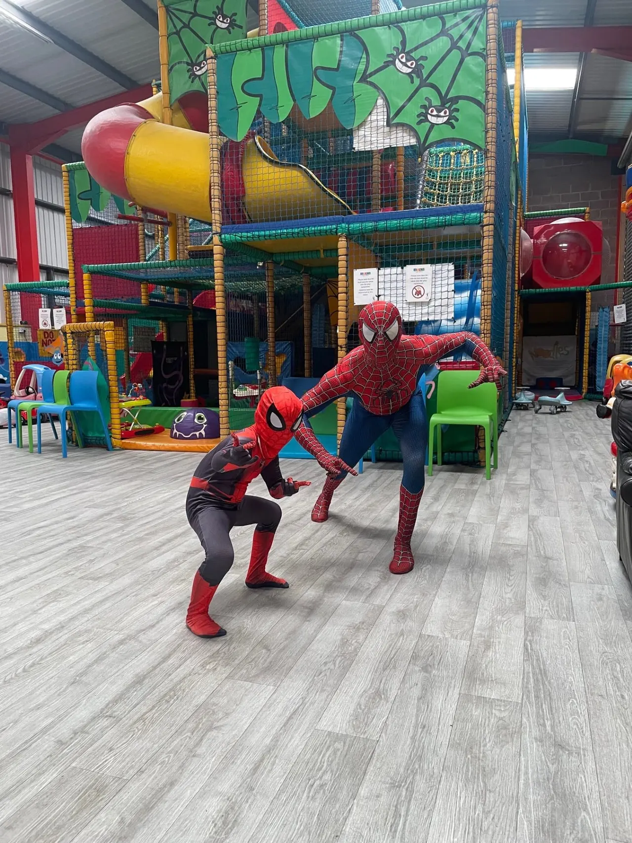 Two people in Spider-Man costumes pose playfully in front of a large colourful indoor play structure with slides and climbing features.